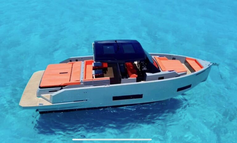 Side profile of 42 ft De Antonio D42 luxury sport yacht charter anchored in crystal-clear turquoise Caribbean, gray hull, orange sun pads, dark blue hardtop, De Antonio logo visible on stern