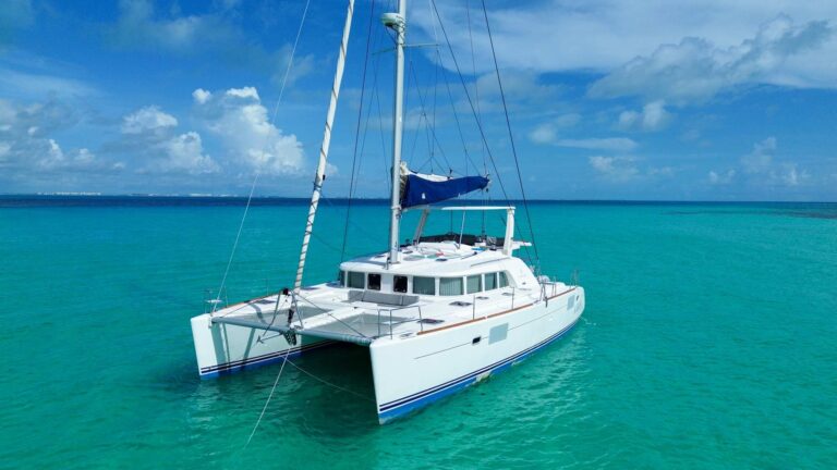 Aerial side view of 50 ft Lagoon catamaran Aarón anchored in flat turquoise Caribbean water, white hull and blue sail cover, Cancun private group charter rental.